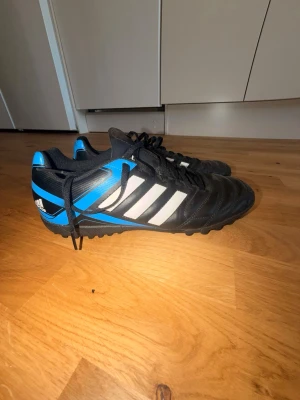 Adidas svart/blå fotbollsskor st: 43 - Säljer ett hardstyle par Adidas fotbollsskor i svart med blå och vita detaljer. Skorna har klassiska tre ränder på sidan, snörning och låg profil. Ovandelen är i syntetmaterial och sulan är mönstrad för bra grepp på planen. Perfekta för dig som vill ha stilrena och funktionella skor.