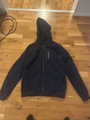 Marinblå hoodie från Sail Racing - Säljer en marinblå hoodie från Sail Racing med dragkedja framtill och en bröstficka med logga och dragkedja. Hoodien har huva med snörning och är tillverkad i ett mjukt material som känns skönt mot huden. Perfekt för dig som gillar sportig och avslappnad stil. Använd sparsamt några gånger 