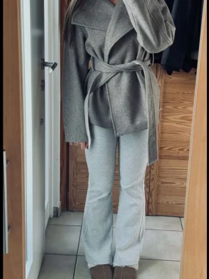 Grå kappa! - Stilren grå kappa från Ella med bred krage och knytskärp i midjan. Kappan har en rak passform och är tillverkad i ett unikt, mjukt tyg som ger en lyxig känsla. Perfekt för att höja vardagslooken med en modern touch.