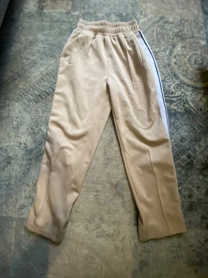 Beige mjukisbyxor med vit sidrand - Säljer ett par beige mjukisbyxor med vit sidrand och elastisk midja. Byxorna har raka ben och en avslappnad passform, perfekta för chill dagar. Materialet känns mjukt och bekvämt mot huden. Snygg detalj med kontrasterande rand längs sidan.