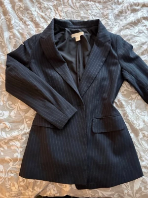 Mörkblå randig kavaj från H&M - Snygg mörkblå kavaj med tunna vita kritstrecksränder från H&M. Klassisk modell med spetsiga slag, två framfickor med lock och helfodrad insida. Perfekt för en stilren och trendig look.