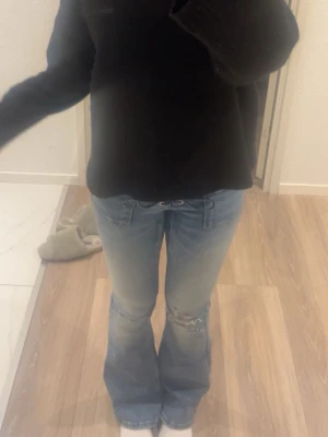 Blå bootcut jeans med snörning - Säljer ett par ljusblå bootcut jeans med snörning i midjan och slitningar vid knäet. Jeansen har en låg midja och klassiska fickor fram och bak. Perfekta för en trendig och avslappnad look.