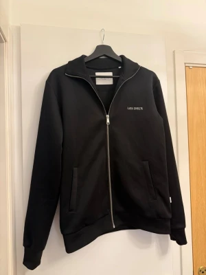 Svart zip hoodie från Les Deux - Snygg svart hoodie från Les Deux med dragkedja framtill och diskret logga på bröstet. Hoodien har två fickor, ribbade muddar och är tillverkad i mjukt material som känns skönt mot huden. Perfekt för en clean och stilren look. Varan är i super skick, inga skador osv. Kostar ca 1199kr ny, detta är gamla modellen