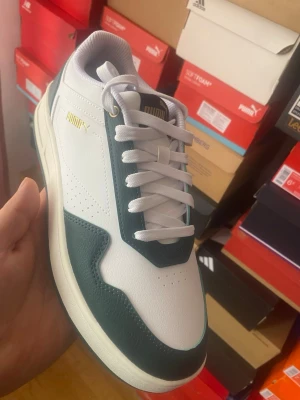 Puma vita och gröna sneakers - Snygga sneakers från Puma i vitt och mörkgrönt med gulddetaljer på sidan och tungan. Skorna har klassisk låg profil, vita skosnören och ovandel i slätt och strukturerat syntetmaterial. Perfekta för dig som gillar stilrena och sportiga sneakers.