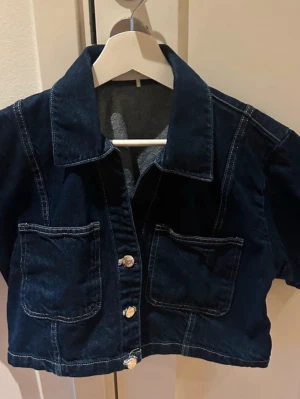 Mörkblå croppad jeansjacka - Snygg mörkblå croppad jeansjacka med stora framfickor och vita kontrastsömmar. Jackan har silverfärgade knappar framtill. 