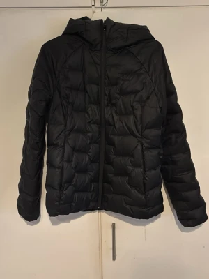 Svart pufferjacka med huva - Snygg svart pufferjacka med huva och dragkedja framtill. Jackan har quiltad design och är vadderad för extra värme. Perfekt för kalla dagar och enkel att matcha med olika outfits. Materialet ser ut att vara syntetiskt och jackan har en modern, något kortare passform.
