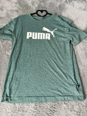 Grön Puma t-shirt med logga - Snygg grön t-shirt från Puma med stor vit logga och text på bröstet. Klassisk rund halsringning och korta ärmar. Tillverkad i mjuk bomullsblandning som känns skön mot huden. Perfekt för en avslappnad och sportig stil.