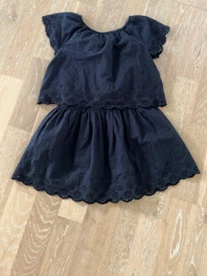 Mörk blå klänning med broderade detaljer - Söt mörk blå kortklänning med broderade blommor längs kanterna och ärmarna. Klänningen har korta ärmar och en lös passform med volangliknande avslut nertill. Perfekt för varma dagar och enkel att styla.