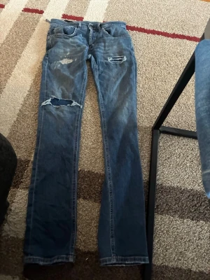 Blå slitna jeans från Dondup - Säljer ett par blå jeans från Dondup med slitningar och hål på låret och knät. Jeansen har klassisk femficksmodell och raka ben. Tillverkade i Italien och har en cool, avslappnad look som passar perfekt till streetwear.modellen heter George slim fit.