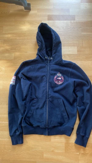 Marinblå hoodie med patchar - Marinblå hoodie med dragkedja och huva. Har patch med amerikansk flagga på ärmen och rund broderad patch på bröstet. Kängurufickor framtill och ribbade muddar. Perfekt för en avslappnad stil.