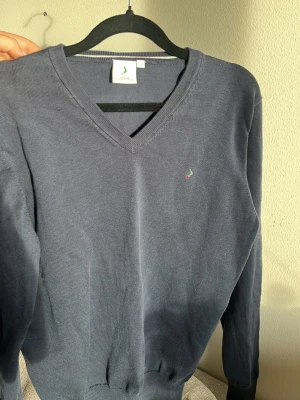 Mörkblå V-ringad tröja  - Mörkblå V-ringad tröja från Boomerang med diskret logga broderad på bröstet. Tröjan har långa ärmar och ribbade muddar vid ärmslut och nederkant. Perfekt för en clean och stilren look.