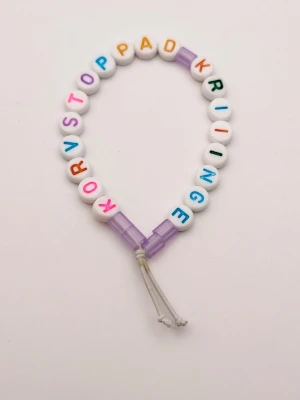 Färgglatt pärlarmband med text - Armband med vita runda pärlor och färgglada bokstäver som bildar orden 'KORVSTOPPAD KRIIINGE'. Armbandet har lila plastdetaljer och justerbar snodd. Perfekt accessoar för dig som gillar unika och personliga smycken.