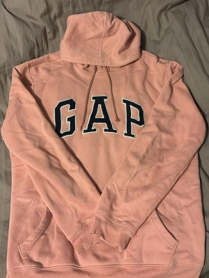 Rosa hoodie från GAP med logga - Säljer en rosa hoodie från GAP med stor broderad logga i svart och vitt på bröstet. Hoodien har huva med dragsko och en stor magficka. Tillverkad i mjuk bomullsblandning, perfekt för chill dagar eller när du vill ha en avslappnad stil.