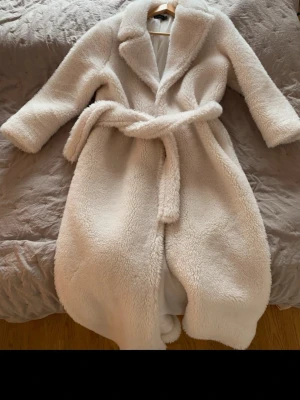 Beige teddyjacka med skärp - Mysig beige teddyjacka med bred krage och knytskärp i midjan. Jackan har en fluffig och mjuk struktur som ger en trendig look. Perfekt för kalla dagar när du vill vara både varm och snygg. Lång modell som ger extra värme.