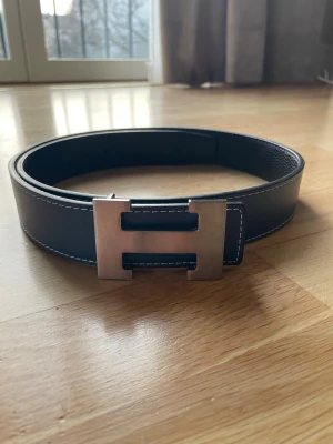 Svart skärp med silverfärgat H-spänne - PRIS KAN DISKUTERAS 🚨Stilrent svart bälte med ett stort, silverfärgat H-format spänne och vita kontrastsömmar längs kanterna. Perfekt accessoar för att lyfta din outfit med en modern touch. Tillverkat i skinn för en exklusiv känsla.