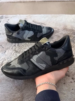 Valentino rockrunner  - Snygga sneakers från Valentino med coolt camo-mönster i svart och grått. Skorna har lågt skaft, svarta skosnören och detaljer i läder och mesh. Perfekt för dig som vill ha en unik och trendig look.