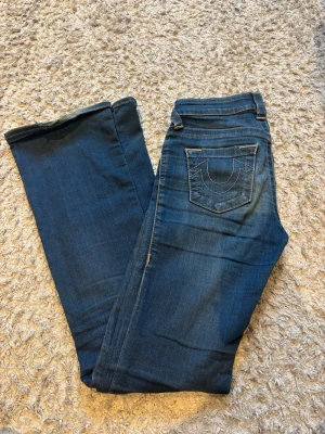 Blå low waist True Religion jeans bootcut - Säljer ett par blå jeans från True Religion med klassisk kontrastsöm och ikonisk fickdesign bak. Modellen har bootcut med låg midja
