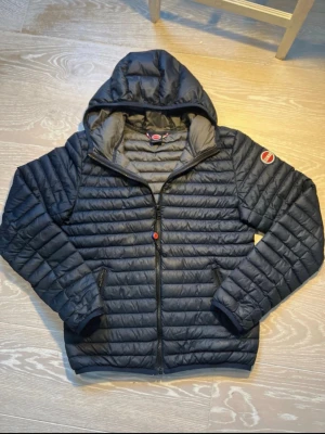 Svart pufferjacka från Colmar - Snygg svart pufferjacka från Colmar med huva och dragkedja framtill. Jackan har två sidofickor med dragkedja och Colmar-logga på ärmen. Quiltad design och lätt vadderad för extra värme. Perfekt för kyliga dagar.