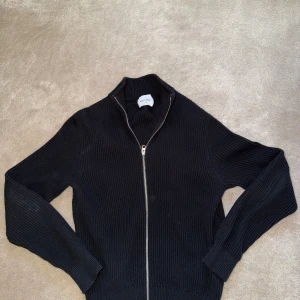 Svart stickad ziptröja från Asket - Säljer en svart stickad ziptröja från Asket med hög krage och hel dragkedja framtill. Tröjan är ribbstickad och har långa ärmar samt en stilren, minimalistisk design. Perfekt för lager på lager och enkel att matcha med olika outfits.