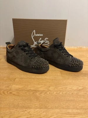 Christian Louboutin svarta spikes sneakers - Grymma svarta sneakers från Christian Louboutin i mocka med ikoniska röda sulor och svarta nitar över tån. Skorna har svarta skosnören och en låg silhuett som ger en edgy vibe. Perfekta för dig som vill sticka ut med exklusiv design och detaljer. Skorna har storlek 41 men passar 42-43