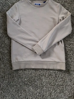 Beige sweatshirt från Jack & Jones - En stilren beige sweatshirt från Jack & Jones med rund halsringning och ribbade muddar vid ärmslut och nederkant. Perfekt basplagg i mjukt material som passar till det mesta. Enkel och clean design utan tryck eller mönster. Den är nästan inte använd alls, jag säljer den bara för att den är lite för liten.