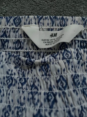 Vit och blå mönstrad kjol H&M - Säljer en vit och blå kjol från H&M med ett snyggt grafiskt mönster. Kjolen har resår i midjan och är gjord i ett lätt och luftigt material, perfekt för varma dagar. Passar dig som gillar en avslappnad men trendig stil.
