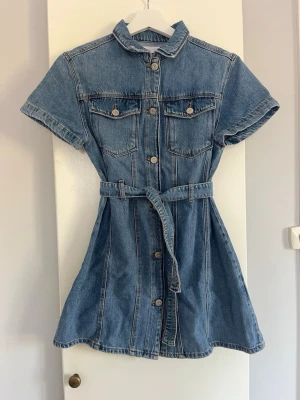 Kort jeansklänning med bälte - Snygg kort jeansklänning i blå denim med knappar framtill, korta ärmar och tillhörande bälte i midjan. Två bröstfickor med lock och krage ger en cool och avslappnad look. Perfekt för dig som gillar en trendig och bekväm stil. 
