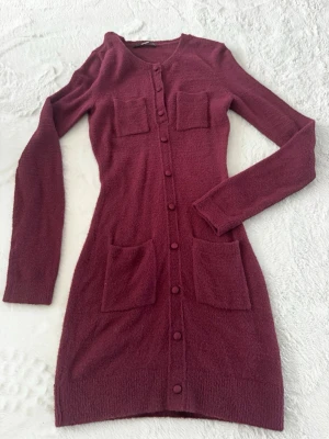 Vinröd stickad långärmad klänning - Vinröd stickad klänning med lång ärm och fyra fickor framtill. Klänningen har knappar längs hela framsidan och en kroppsnära passform. Perfekt för dig som gillar en enkel men snygg look.