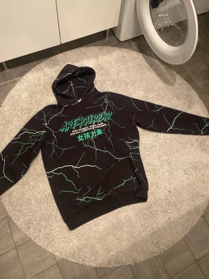 Svart hoodie med grön text och blixtar - Svart hoodie från AreYouHype med grön och vit blixtdesign över hela tröjan. Tryck på bröstet med texten 'AREYOUHYPE' och citat samt japanska tecken. Huva och magficka framtill. Perfekt för dig som gillar streetwear och unika prints.