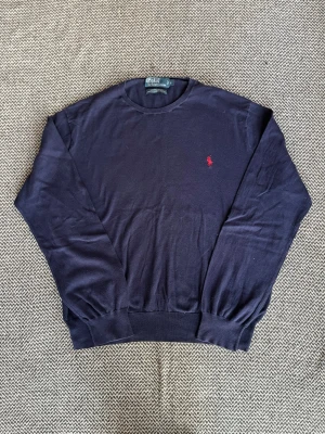 Mörkblå tröja från Polo Ralph Lauren - Marinblå Polo Ralph Lauren tröja med klassisk röd broderad logga på bröstet. Tröjan har rund halsringning, långa ärmar och ribbade muddar vid ärmslut och nederkant. Perfekt för lager på lager och stilren look. 100% pima bomull. Mycket bra skick, inga defekter förutom lappen är lös i ett hörn (väldigt lätt lagning). Storlek L. Axelbredd: 51 cm Längd: 66 cm