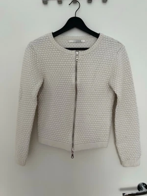 Vit strukturerad kofta med dragkedja - Snygg vit kofta från Sibin Linnebjerg med rund halsringning och strukturstickning. Koftan har en silverfärgad dragkedja framtill och ribbade muddar vid ärmslut och nederkant. Perfekt att slänga över en topp för en clean look.