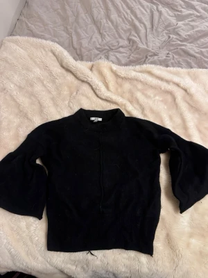 Svart stickad tröja med vida ärmar Dagmar - En svart stickad tröja med rund hals och vida, trumpetformade ärmar. Tröjan har en enkel design utan synliga mönster och är tillverkad i ett mjukt stickat material. Perfekt för dig som gillar en stilren och trendig look. Från DAGMAR