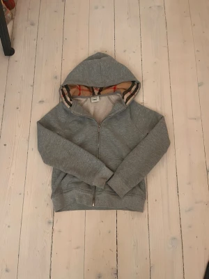 Grå zip-hoodie från Burberry - Snygg grå zip-hoodie från Burberry med klassiskt rutigt foder i huvan. Tillverkad i mjukt bomullsmaterial och har dragkedja framtill samt ribbade muddar. Perfekt för en avslappnad och trendig look.