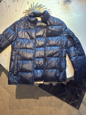 Blå dunjacka från Dolomite - Snygg blå dunjacka från Dolomite med quiltad design och hög krage. Jackan har dragkedja framtill och två sidofickor. På ärmen sitter ett Dolomite-märke med italienska flaggans färger. Perfekt för kalla vinterdagar.