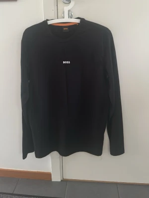 Svart långärmad topp från BOSS - Svart långärmad topp från BOSS med enkel och stilren design. Toppen har rund halsringning och en liten vit BOSS-logga på bröstet. Perfekt för en clean och klassisk look.