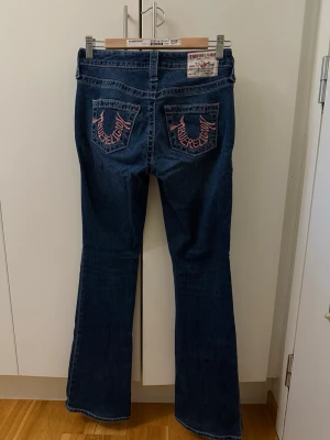 True Religion bootcut jeans mörkblå - Mörkblå bootcut jeans från True Religion med klassiska kontrastsömmar och broderade bakfickor i rosa. Jeansen har fem fickor, låg midja och stängs med knapp och dragkedja. Snygg passform och ikonisk logga på bakfickorna.