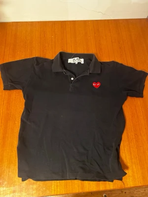 Svart pikétröja Comme des Garçons Play - Svart pikétröja från Comme des Garçons Play med klassisk krage och knappar framtill. Tröjan har det ikoniska röda hjärta-märket broderat på bröstet. Tillverkad i bomull och har korta ärmar. Perfekt för en avslappnad men stilsäker look. Priset kan diskuteras.