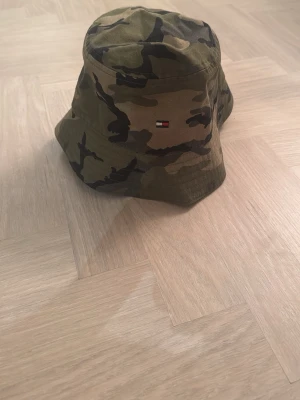 Camo bucket hat från Tommy Hilfiger - Säljer en bucket hat i camomönster från Tommy Hilfiger. Hatten har klassisk form. På sidan sitter det ikoniska Hilfiger-flaggan broderad. Perfekt för dig som vill ha en streetwear-vibe.