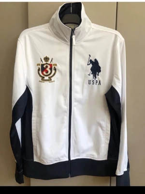 Vit USPA ziptröja med brodyr - Snygg vit ziptröja från US Polo Assn med mörkblå detaljer på sidor och ärmar. Tröjan har hög krage, dragkedja framtill och broderade loggor på bröstet och ärmen. Perfekt för en sportig och stilren look.