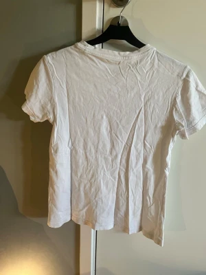 Vit Adidas t-shirt med rosa logga - Säljer en klassisk vit t-shirt från Adidas med stor rosa Trefoil-logga och text på bröstet. T-shirten har rund halsringning och korta ärmar. Tillverkad i mjuk bomull för skön känsla och enkel stil.