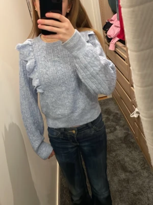 Ljusblå stickad tröja med volanger - Ljusblå stickad tröja med snygga volanger längs axlarna och ett diskret mönster i stickningen. Tröjan har rund hals och långa ärmar med ribbade muddar. Perfekt för dig som gillar en söt och trendig look.