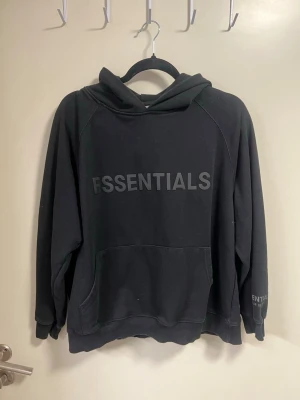 Svart Essentials Fear of God hoodie - Svart hoodie från Essentials Fear of God med stor logga på bröstet och mindre tryck på ärmen. Klassisk känguruficka framtill och huva. Tillverkad i mjukt bomullsmaterial som känns skönt mot huden. Perfekt för en avslappnad och trendig streetwear-look.