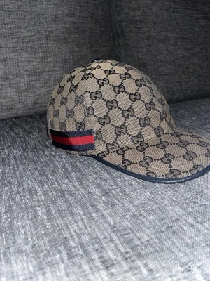 Gucci keps med GG-mönster - Snygg beige keps från Gucci med klassiskt GG-monogram över hela kepsen och en ikonisk röd och marinblå rand på sidan. Kepsen har böjd skärm och justerbar rem bak. Perfekt för dig som vill sticka ut med en lyxig accessoar.