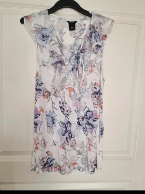 Blommig vit linnetopp med knyt - Supersöt ärmlös linnetopp i vitt med blommigt mönster i lila, blått och korall. Toppen har knytning vid halsen och en lätt, luftig känsla – perfekt för varma dagar. Snygg som diskrer färgklick under kavaj eller ensam till vita byxor tex.