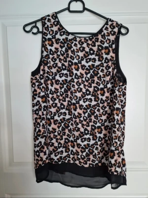Leopardmönstrad ärmlös topp - Snygg ärmlös topp med leopardmönster i svart, brun och beige. Toppen har rund halsringning och transparent svart kant nertill samt öppen ryggdetalj. Perfekt för dig som vill sticka ut med ett djurmönster och en cool twist baktill.