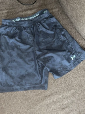 Under Armour träningsshorts blå - Snygga blå träningsshorts från Under Armour med diskret camouflagemönster och elastisk midja med snörning. Perfekta för gymmet eller löprundan, tillverkade i lätt och snabbtorkande polyester. Logga på benet för sportig look. Passar s-M
