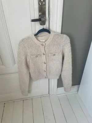 Beige fluffig kofta med fickor - Mysig beige kofta i fluffigt material med rund halsringning och dekorativa bröstfickor. Koftan har långa ärmar, ribbade muddar och knappar framtill. Perfekt för lager på lager och en avslappnad form💕