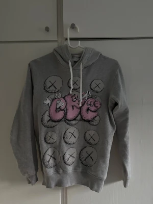 CDG hoodie - Säljer en grå hoodie med huva och vita dragsnören. På framsidan finns ett stort grafiskt tryck med rosa och vita bokstäver samt cirklar med kryss. Hoodien har ribbade muddar vid ärmar och nederkant. Perfekt för dig som gillar streetwear och coola prints. Passar slim fit storlek XS kvitto finns