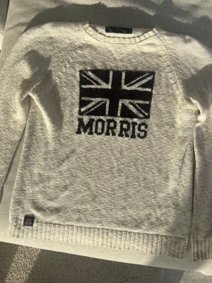 Vit stickad tröja från Morris - Vit stickad tröja från Morris med svart Union Jack-flagga och texten 'MORRIS' framtill. Tröjan har rund halsringning, ribbade muddar och en liten patch nertill. Perfekt för dig som gillar klassisk och stilren design.