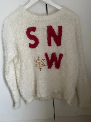 Vit fluffig stickad SNOW-tröja - Vit stickad tröja med fluffig känsla och röda bokstäver som bildar ordet 'SNOW' på framsidan. Under bokstäverna finns en glittrig snöflinga i guldpaljetter. Perfekt för vintern och julen.